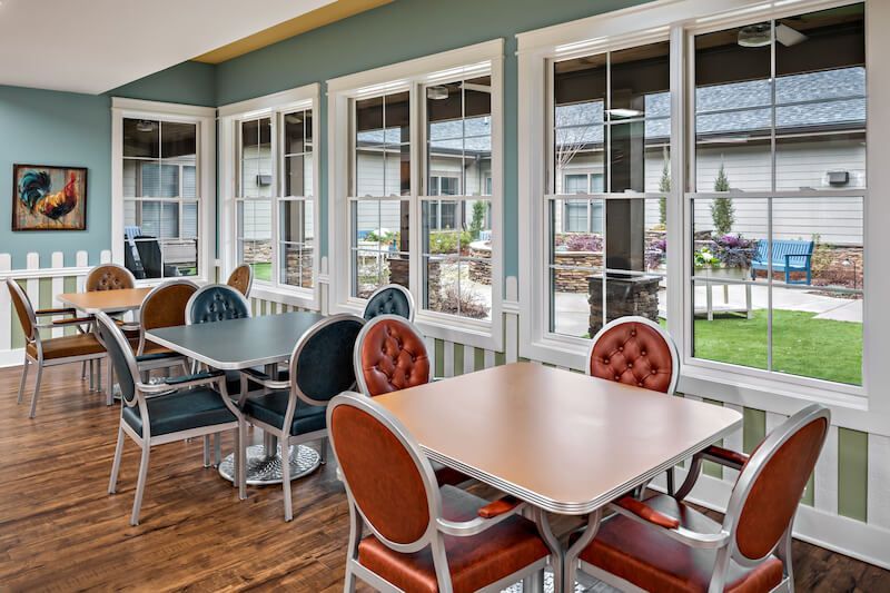 A bright dining room with tables and chairs, large windows overlooking a green yard.