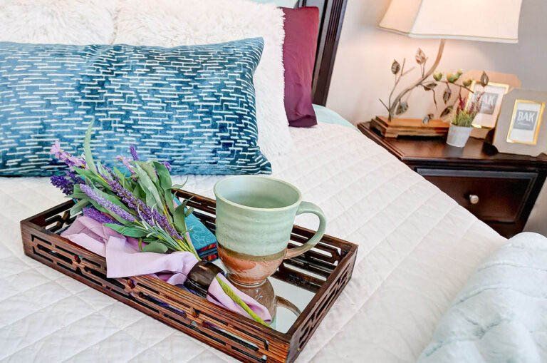 Bed with a tray holding a mug, flowers, and napkins. A nightstand with a lamp and photos is nearby.