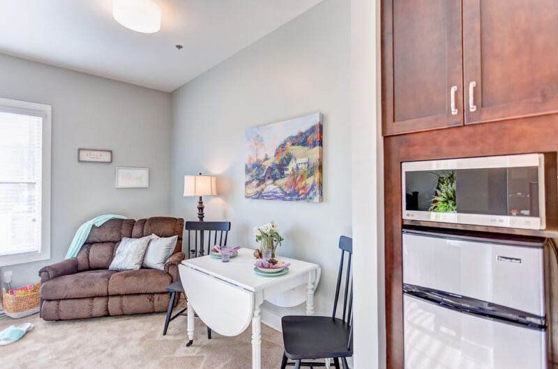 Small living space: Brown recliner, white table with two chairs, microwave above a mini-fridge, and wall art.