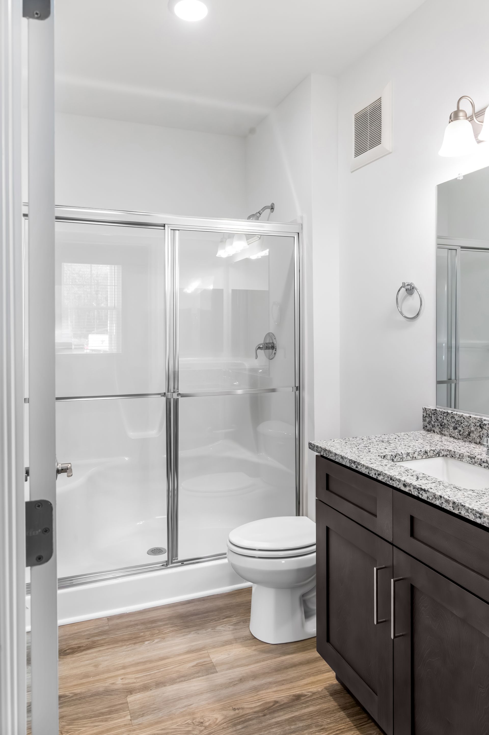 Bathroom with a shower, toilet, and vanity with granite countertop. White walls, brown wood-look flooring.