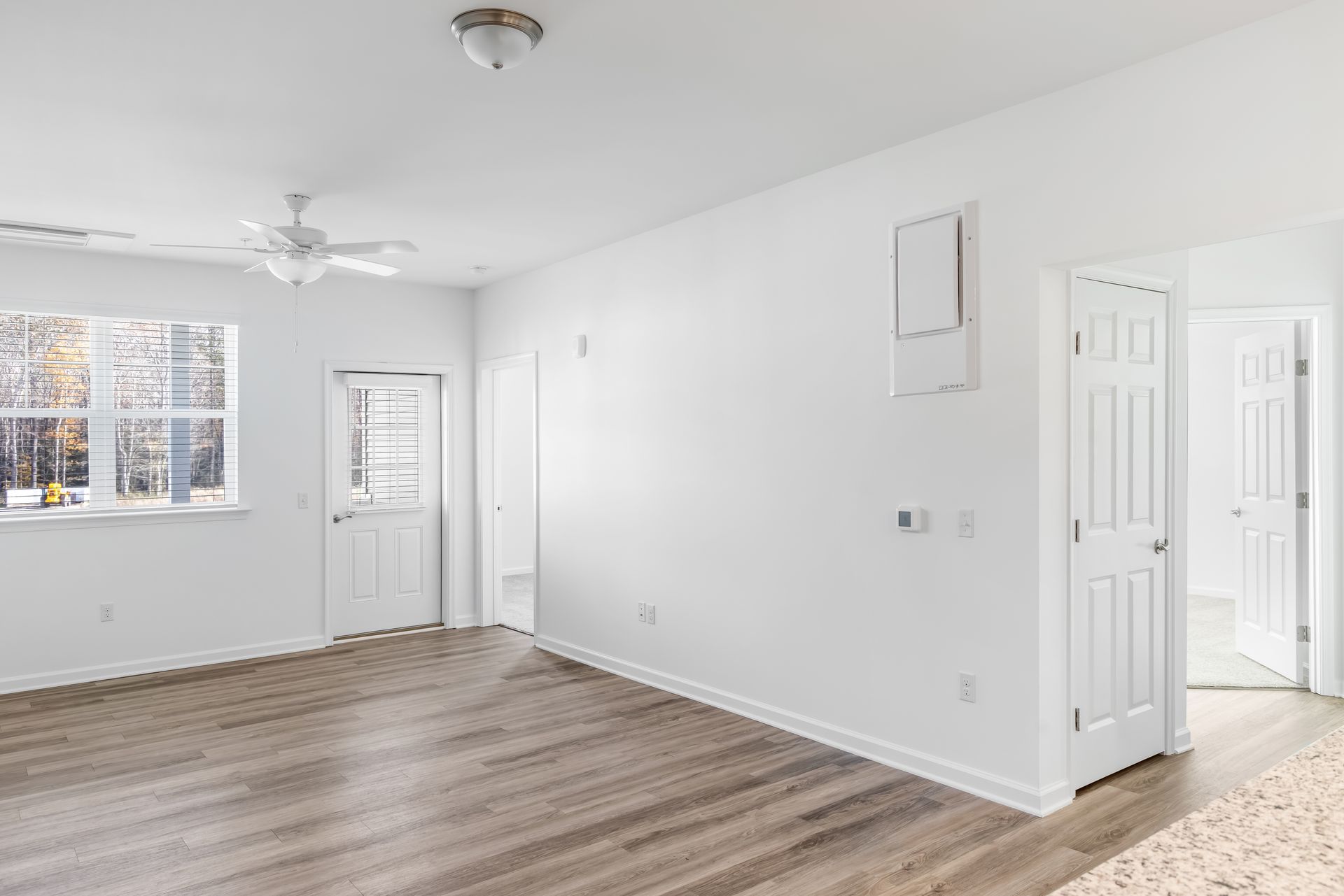 Bright, empty room with white walls, doors, and a ceiling fan. Light wood-look flooring. Window on the left.