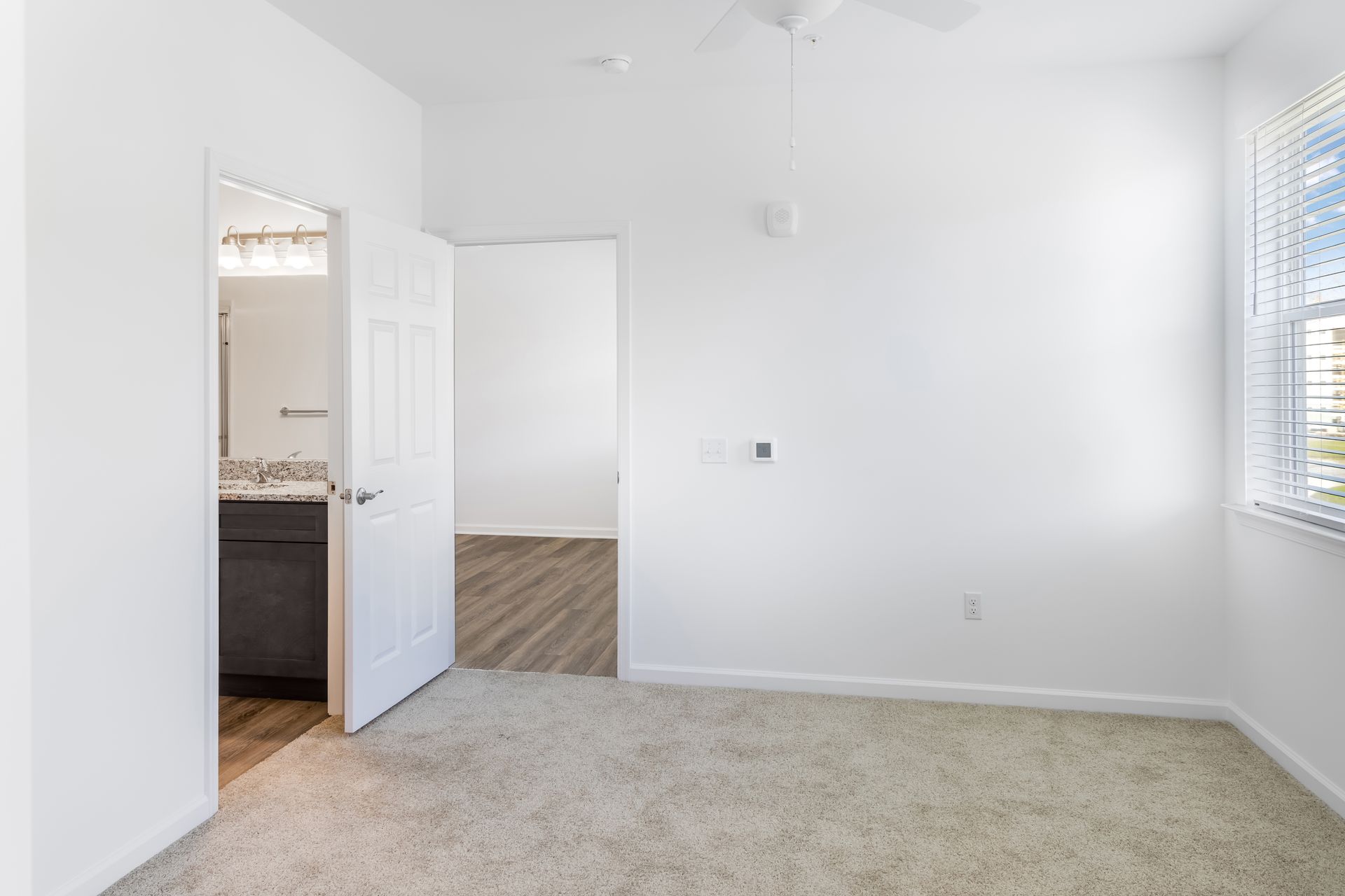 Empty bedroom with tan carpet, white walls, and a doorway to a bathroom.