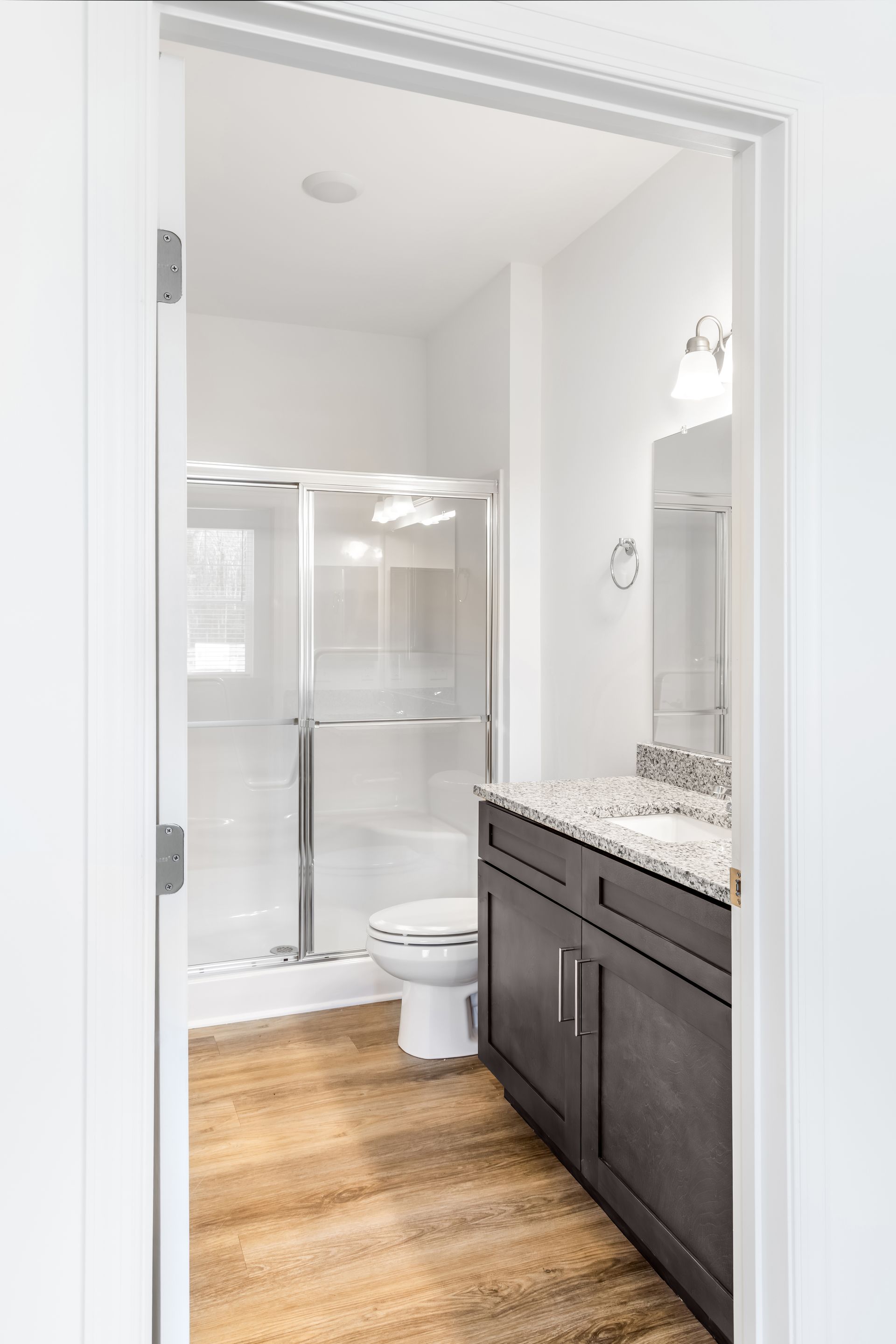 Bathroom with shower, toilet, vanity, and mirror visible through a doorway. Brown wood-look floor, white walls.