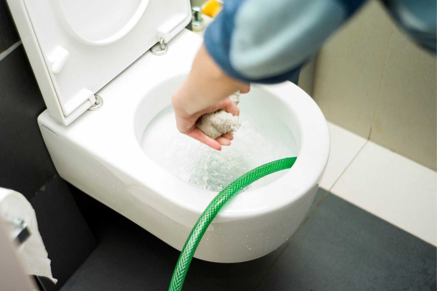 A person's hand holds a cloth in a toilet bowl while a green hose pours water.