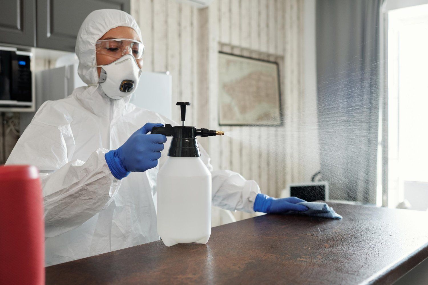 Person in protective gear spraying disinfectant on a countertop in a kitchen.