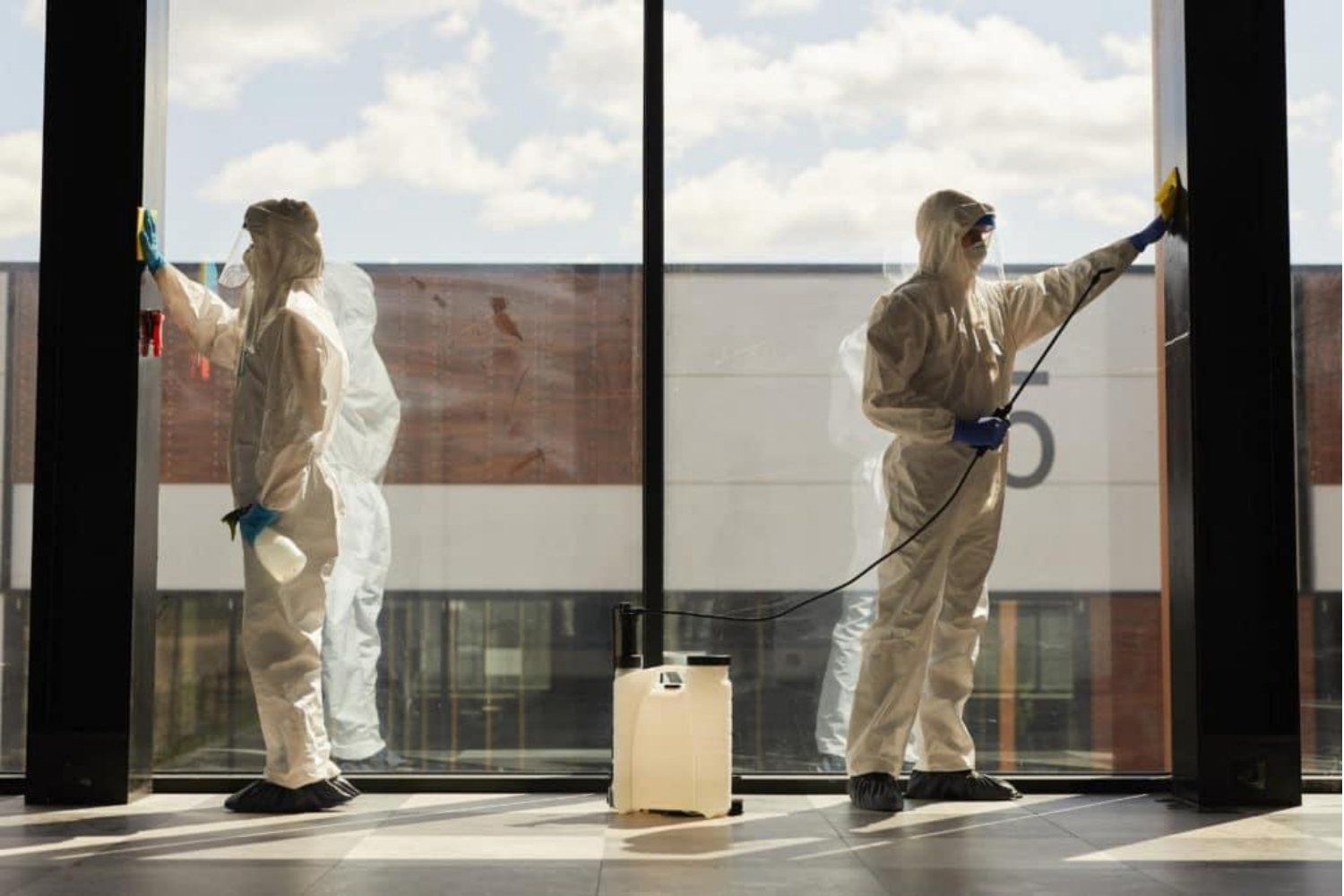 Two people in hazmat suits cleaning a large window, one spraying disinfectant, the other wiping. Inside a building.