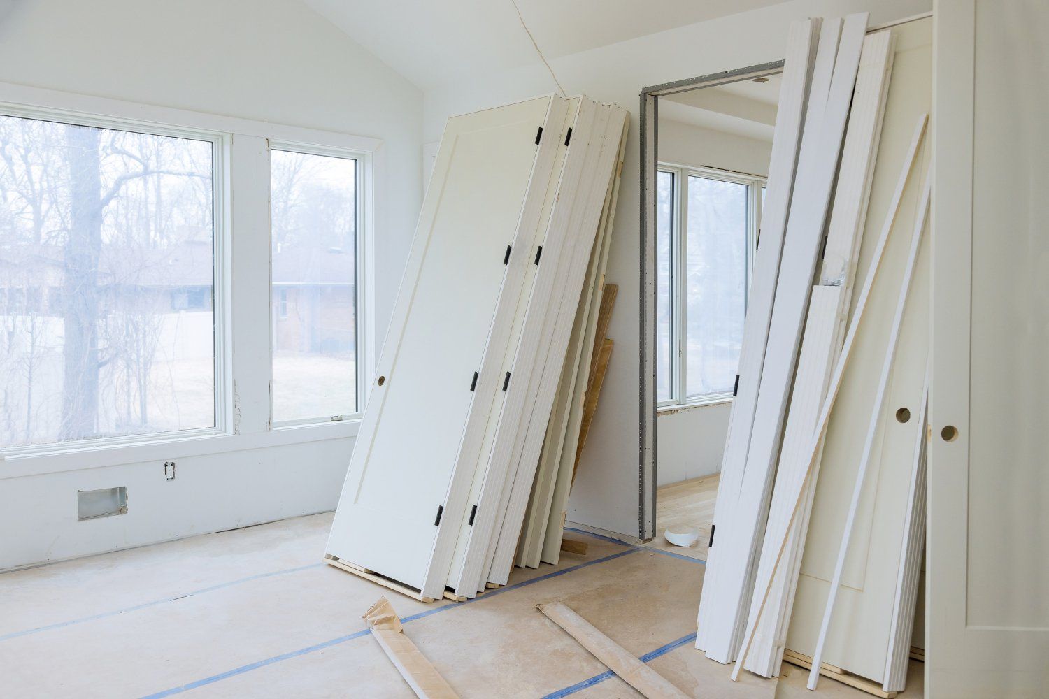 Interior room under renovation with a stack of white doors and trim leaning against the wall near a doorway and window.