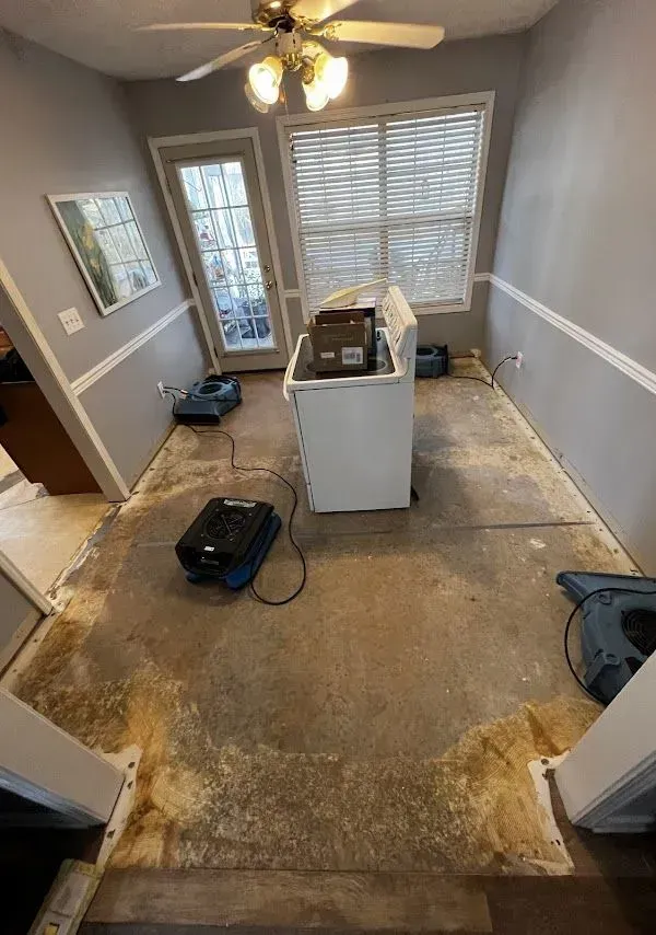 A room with exposed concrete floor, drywall wainscoting, and a washing machine. Dehumidifiers and fans are drying the space.