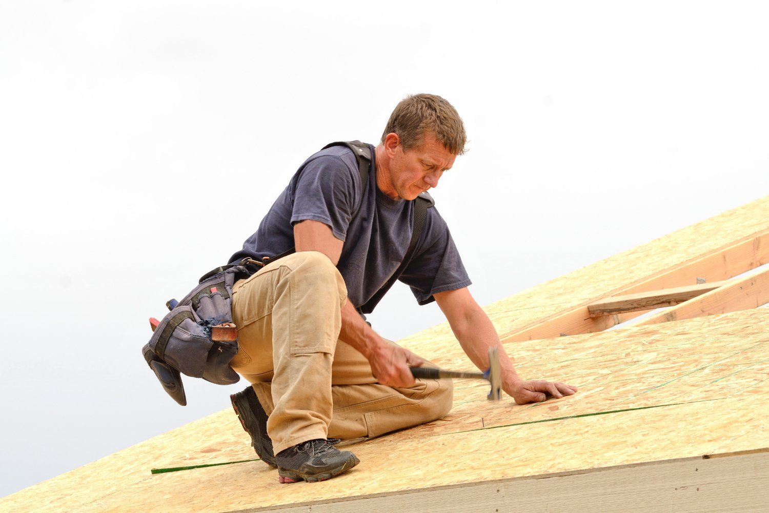 Roofer kneels on a wooden roof, hammering a nail. He wears work clothes and a tool belt under a cloudy sky.