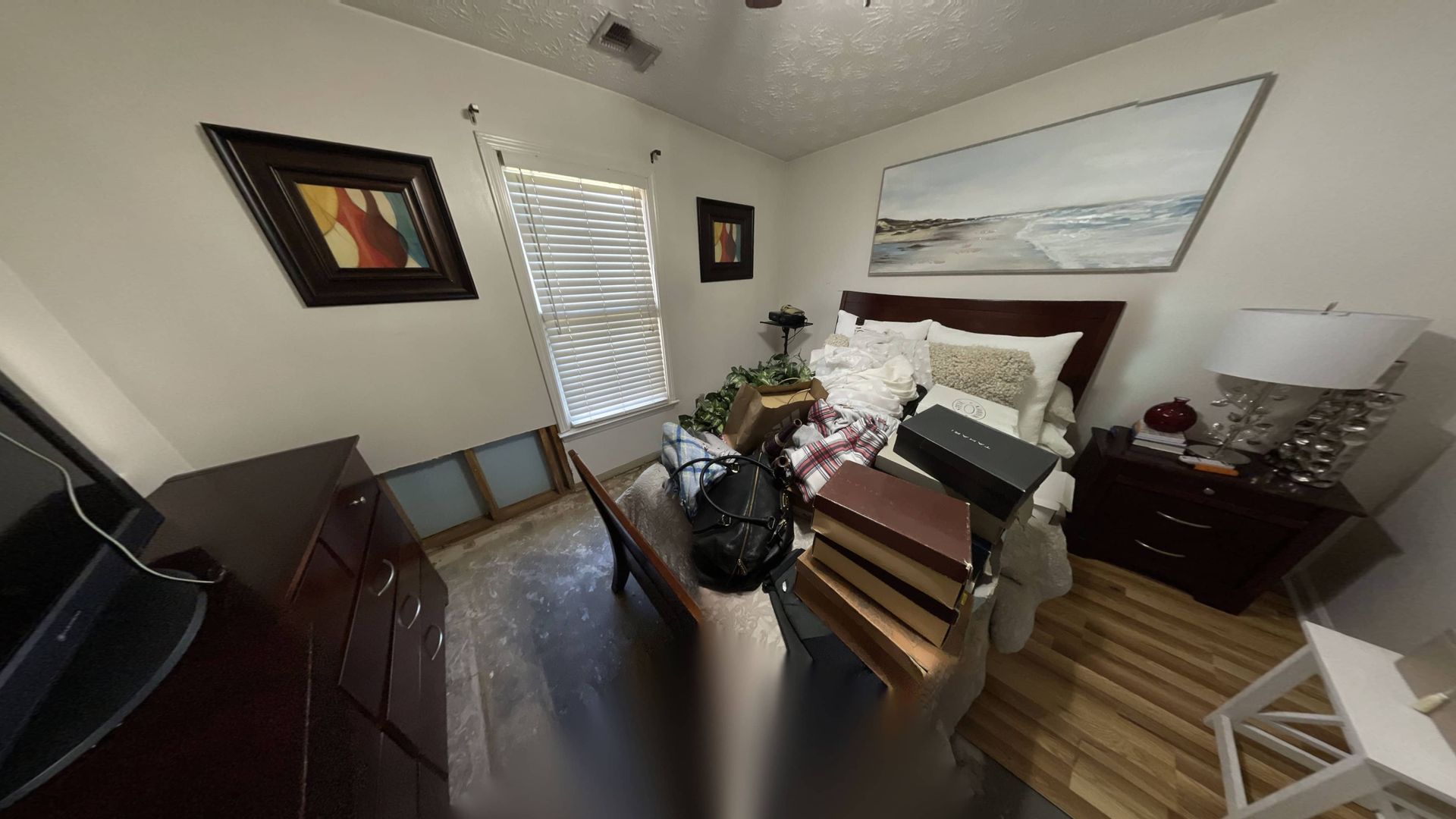 Bedroom with a dresser, bed, nightstand, and artwork on the walls. The bed has pillows and a blanket, and the room appears cluttered.