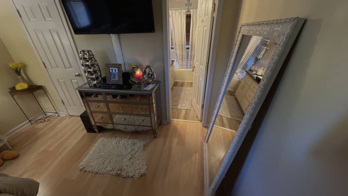 A narrow hallway with a mirror, dresser, rug, and TV. Light-colored wood floors and walls, with a view toward a distant doorway.