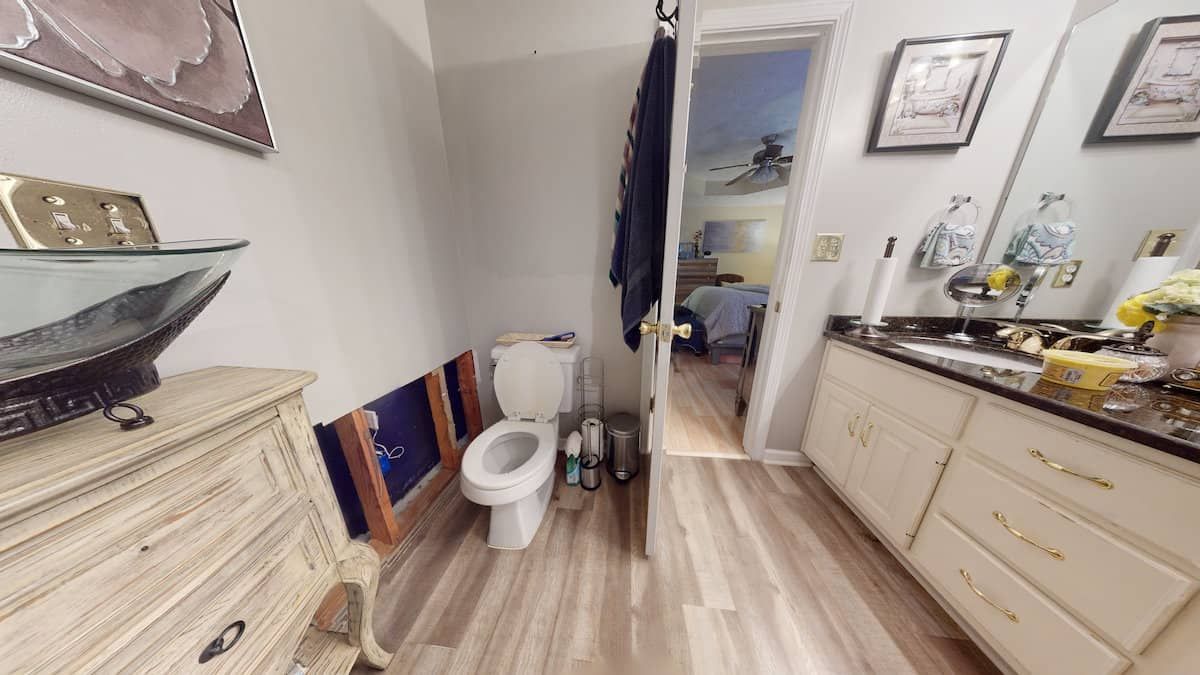 Bathroom with white toilet, vanity, and partially damaged wall. Light wood-look flooring and a doorway to a bedroom are visible.