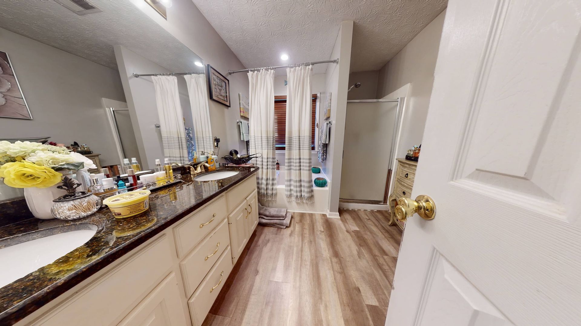 Bathroom with a double vanity, shower stall, and bathtub. Features light wood-look flooring, white cabinets, and a shower curtain.