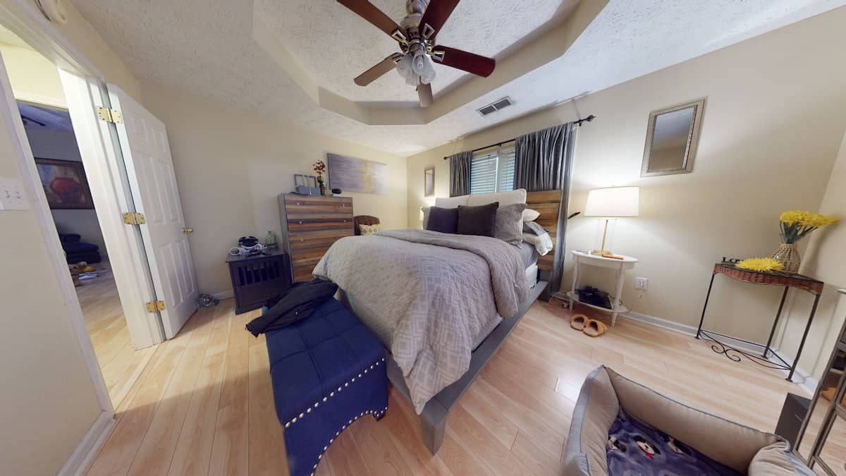 Bedroom with a bed, dresser, nightstands, and a chair. Light-colored wood floors, walls, and ceiling with a ceiling fan.