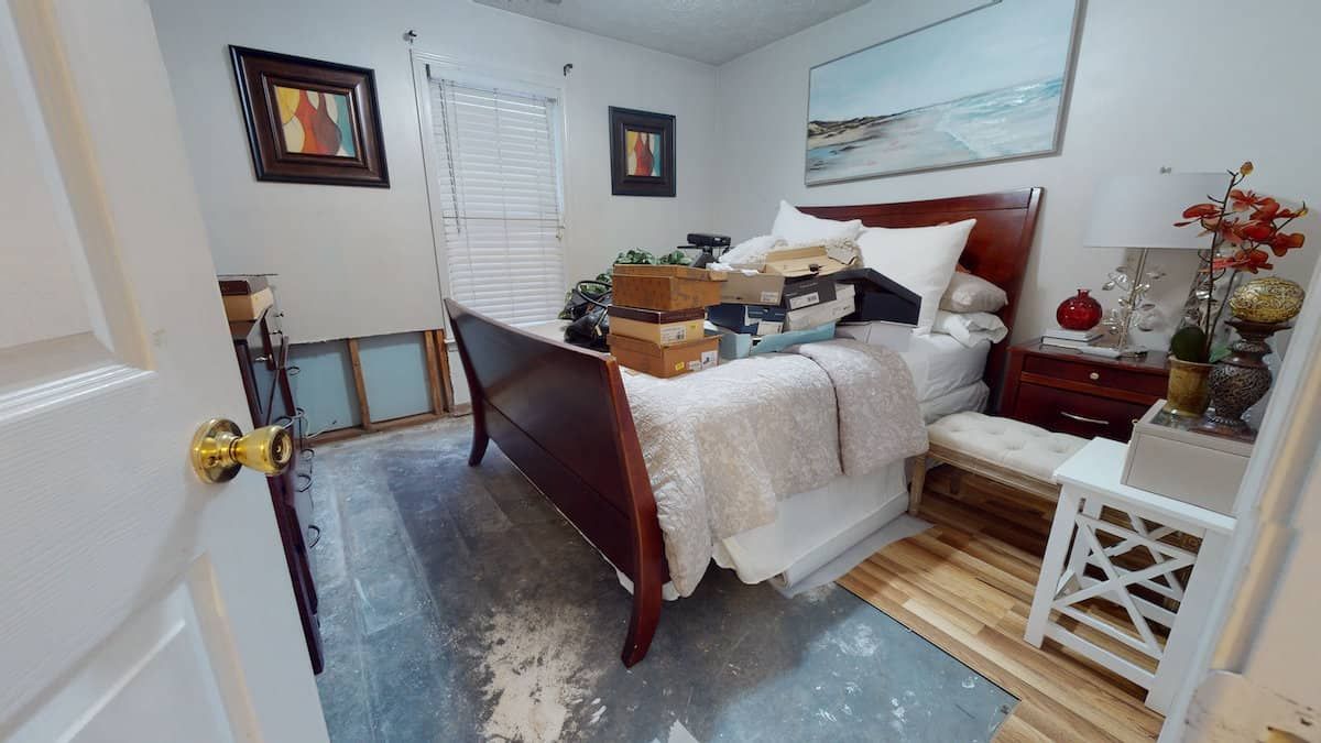 Bedroom interior with a bed, nightstand, and various items scattered around. The bed is cluttered. The room has art and a window.