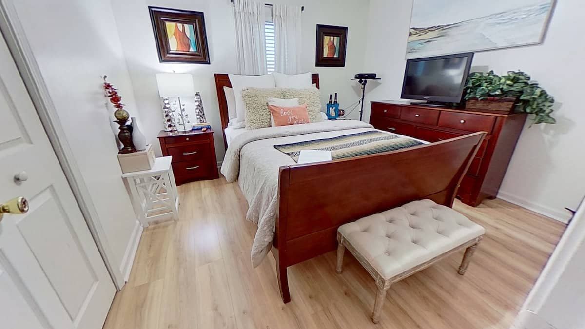 Bedroom with a wooden bed, dresser, and nightstand. Bed has decorative pillows and a quilt. Light-colored flooring and walls.