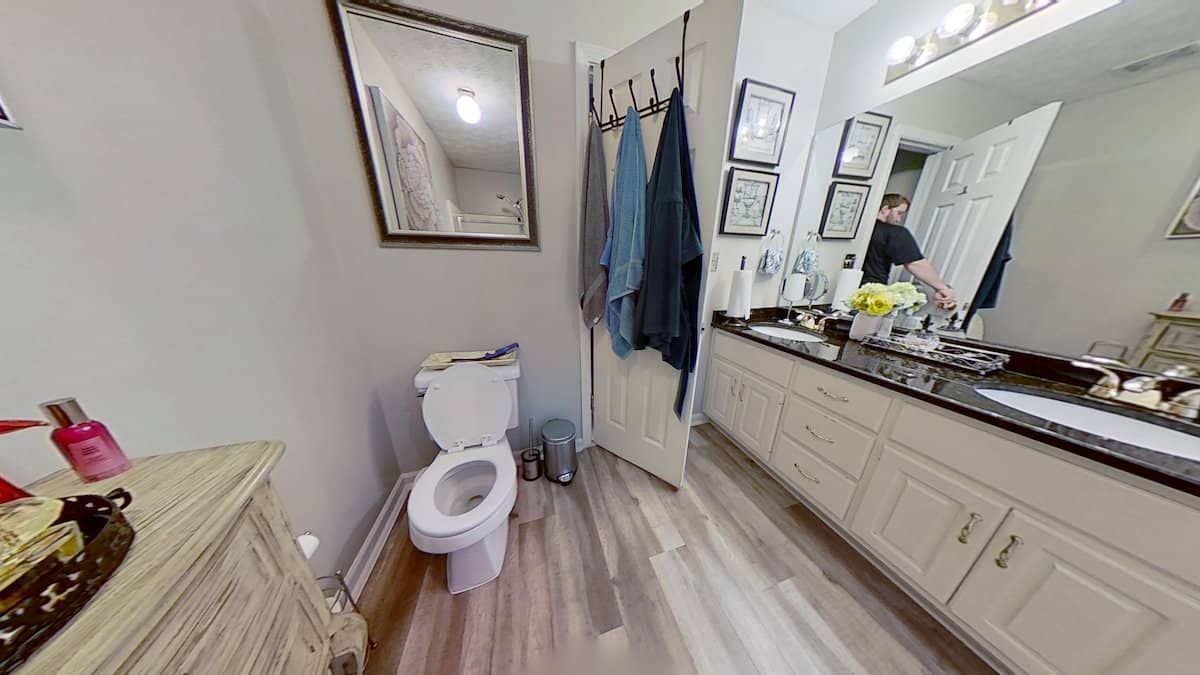 Bathroom with a toilet, vanity, and mirror. The floor is light brown, and there are towels hanging on the door.