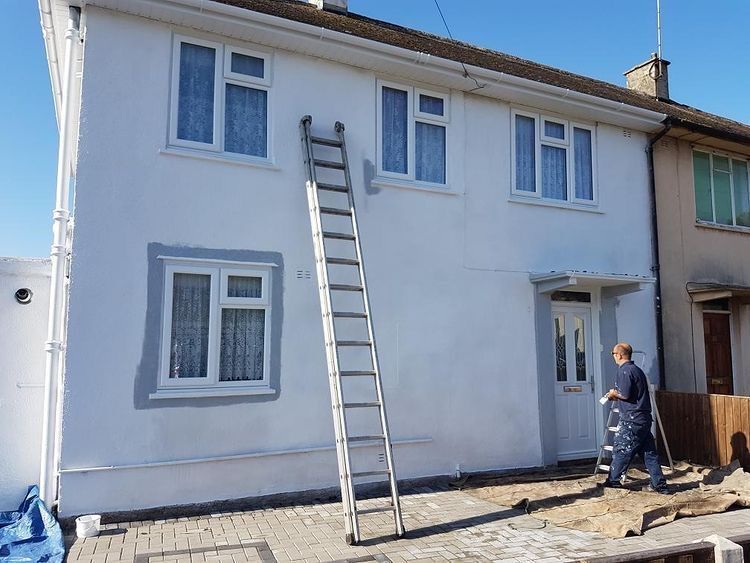 A house being painted white with a ladder propped against it. A person is painting near the door.