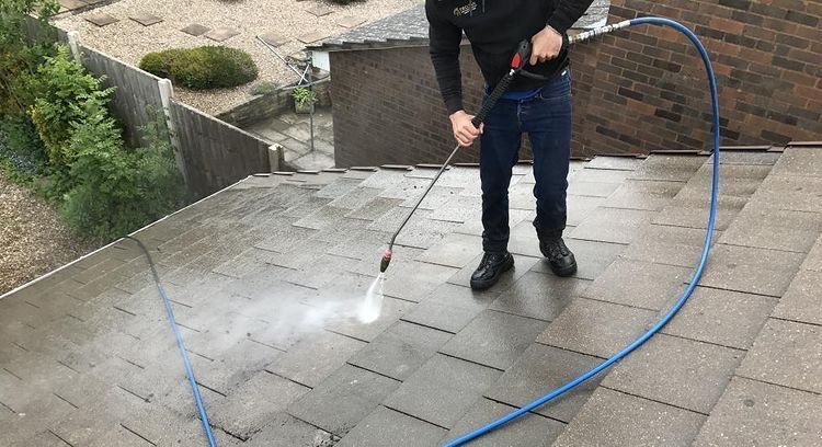 A person power washing a residential tiled roof with a blue hose on a cloudy day.