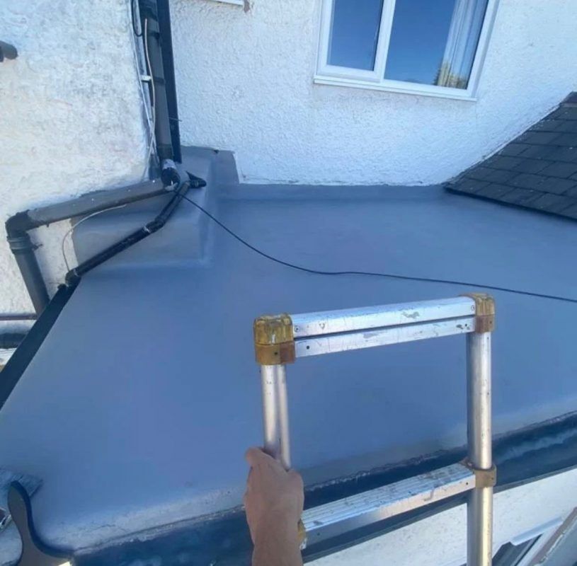 A person holding a metal ladder while standing on a newly coated, grey flat roof next to a house wall and tiled roof.