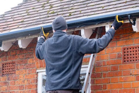 Person on ladder attaching gutters to a roof on a brick house.