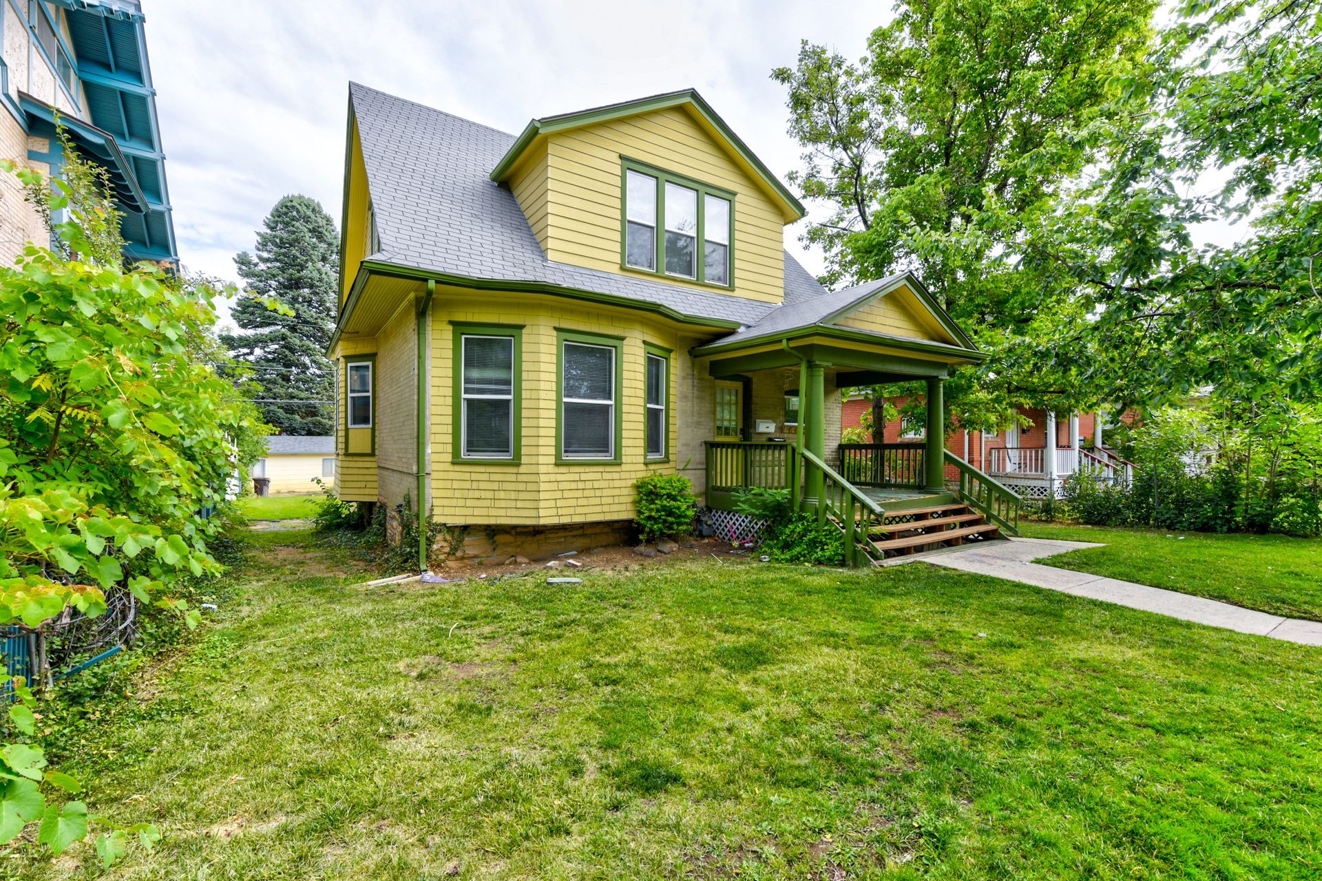 Yellow house with green trim and porch; grassy yard, sidewalk.