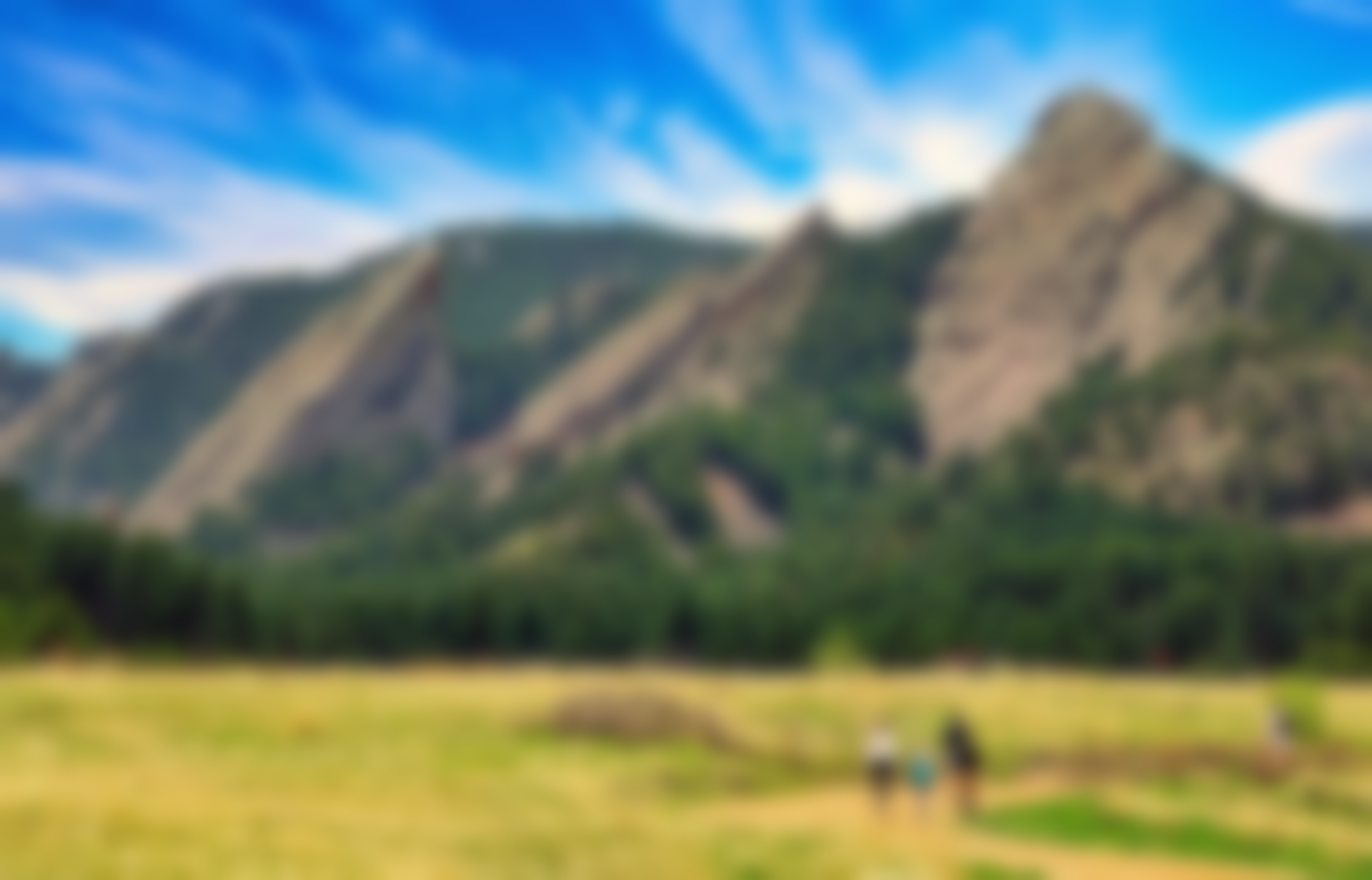 Mountains with a field and three blurred figures walking on a trail under a blue sky.