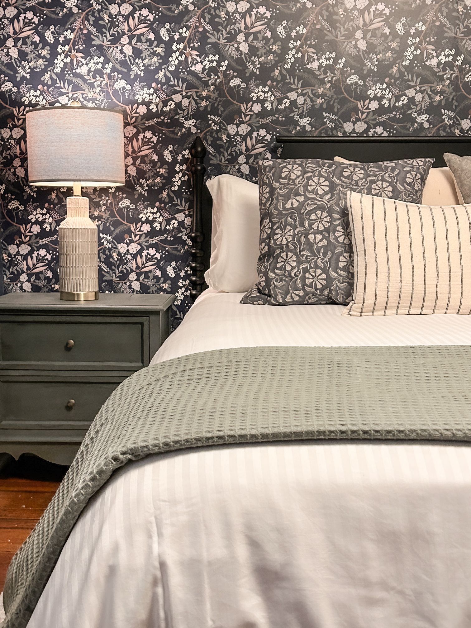 A bedroom with a bed , nightstand , lamp and floral wallpaper.