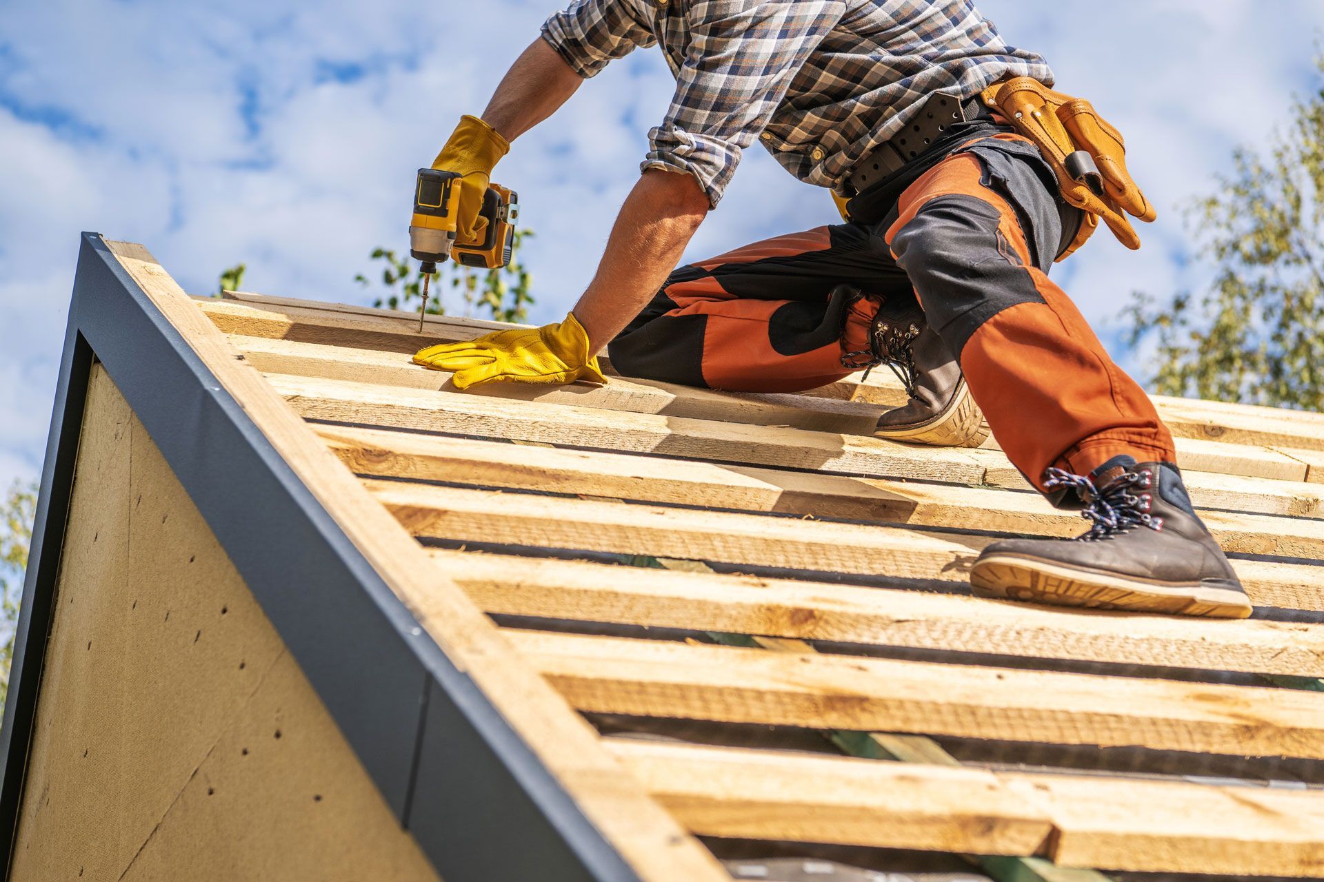 Roofer wearing yellow gloves and safety gear working on a roof with a power drill on a sunny day.