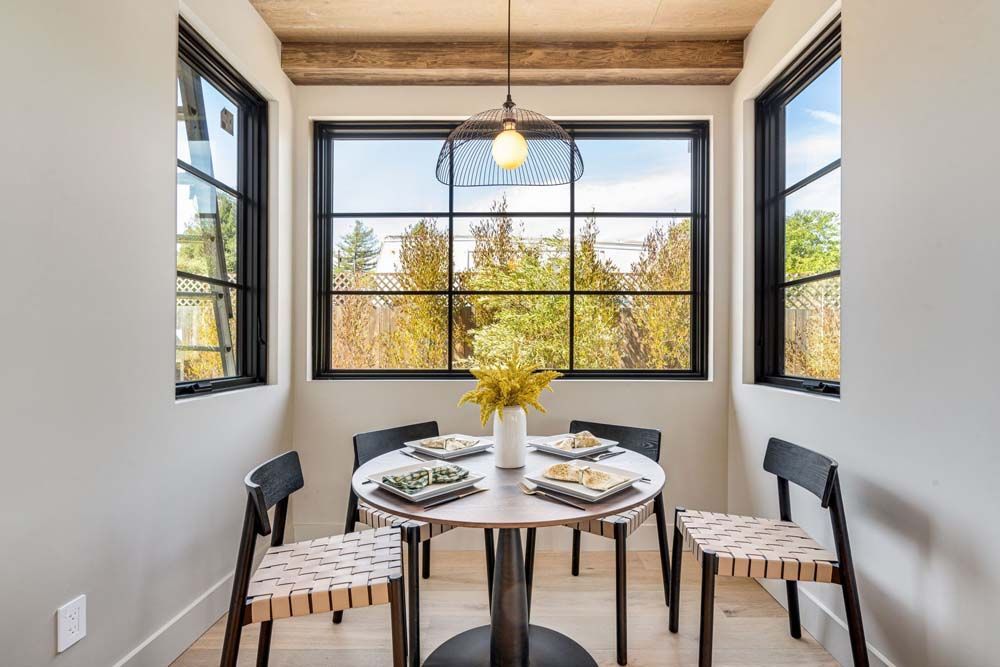 Dining nook with round table and chairs, large windows with a view of trees, and a hanging light.