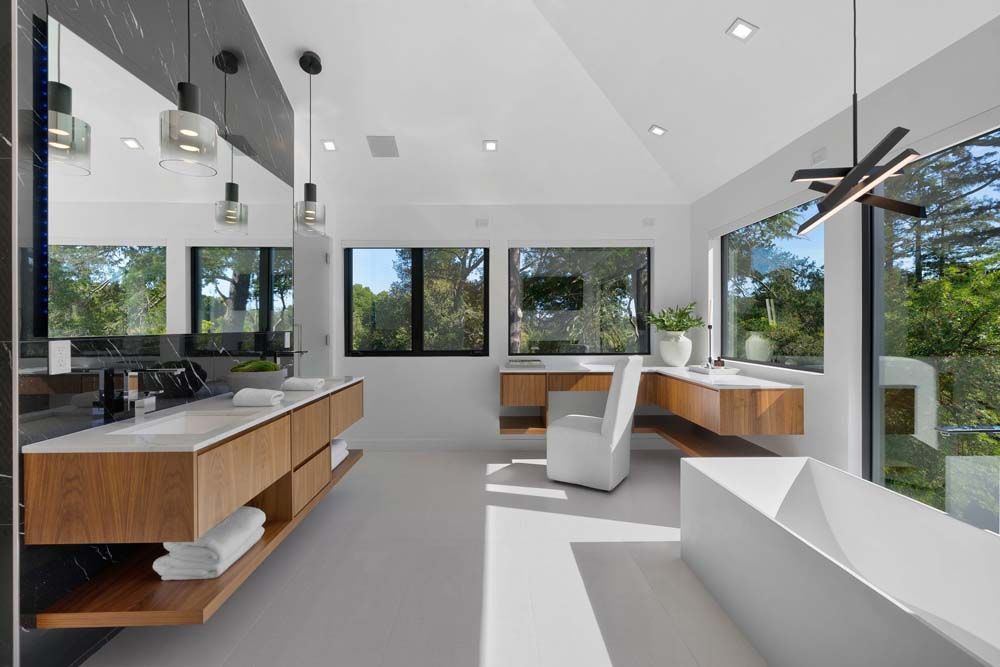 Modern bathroom with floating wooden vanities, white walls, and large windows overlooking trees.
