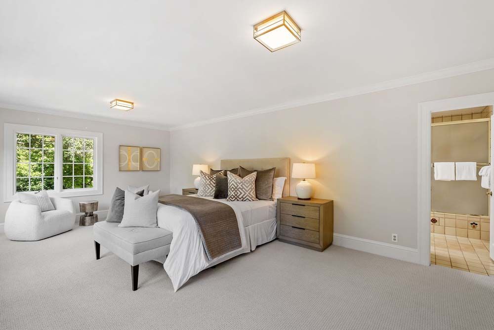 Bedroom with light walls, bed, seating area, window, and doorway to a bathroom.