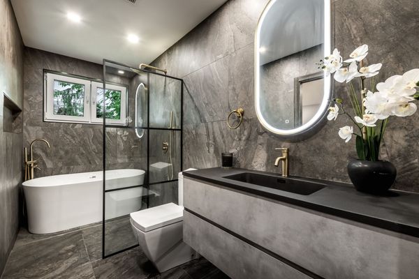 Modern bathroom with gray stone walls, glass shower, freestanding tub, and illuminated oval mirror.