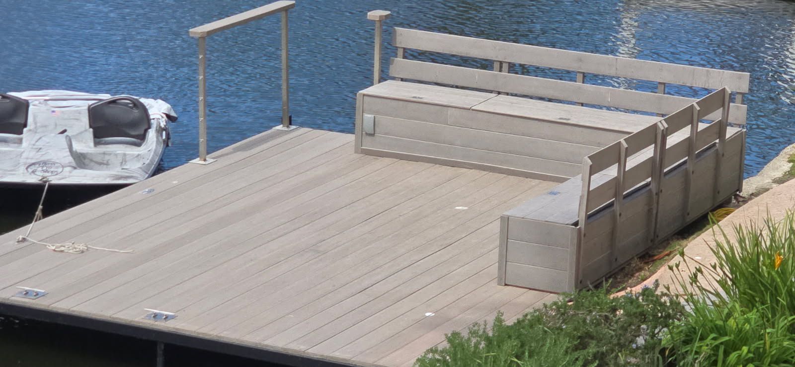 A light-gray wooden dock sits on calm water, featuring an L-shaped bench and a small boat moored to the side.