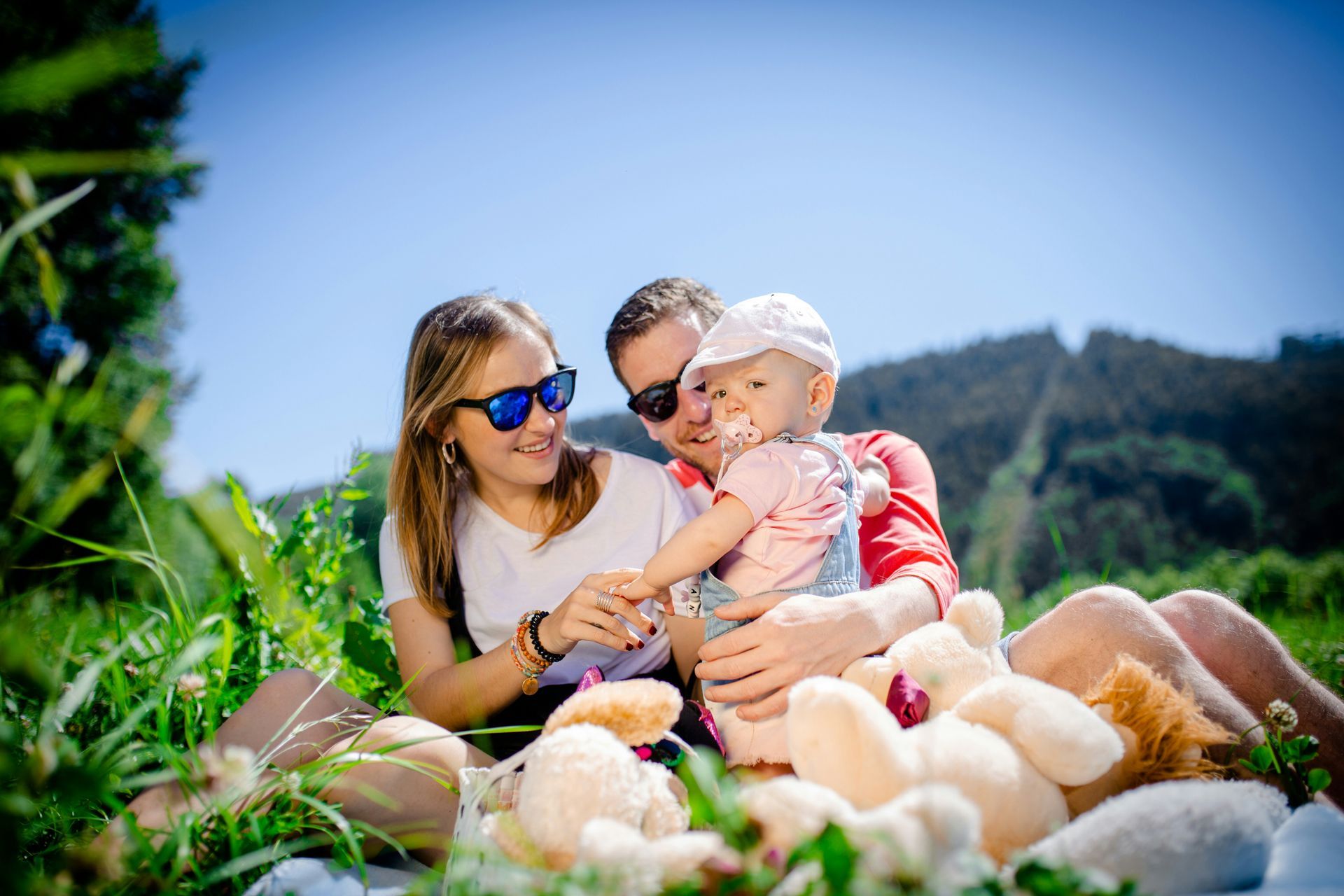 A family is sitting on a blanket in the grass holding a baby and a teddy bear.
