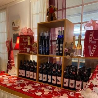 A Valentine’s display of wine bottles in wooden shelves on a heart-patterned table, decorated with teddy bears.