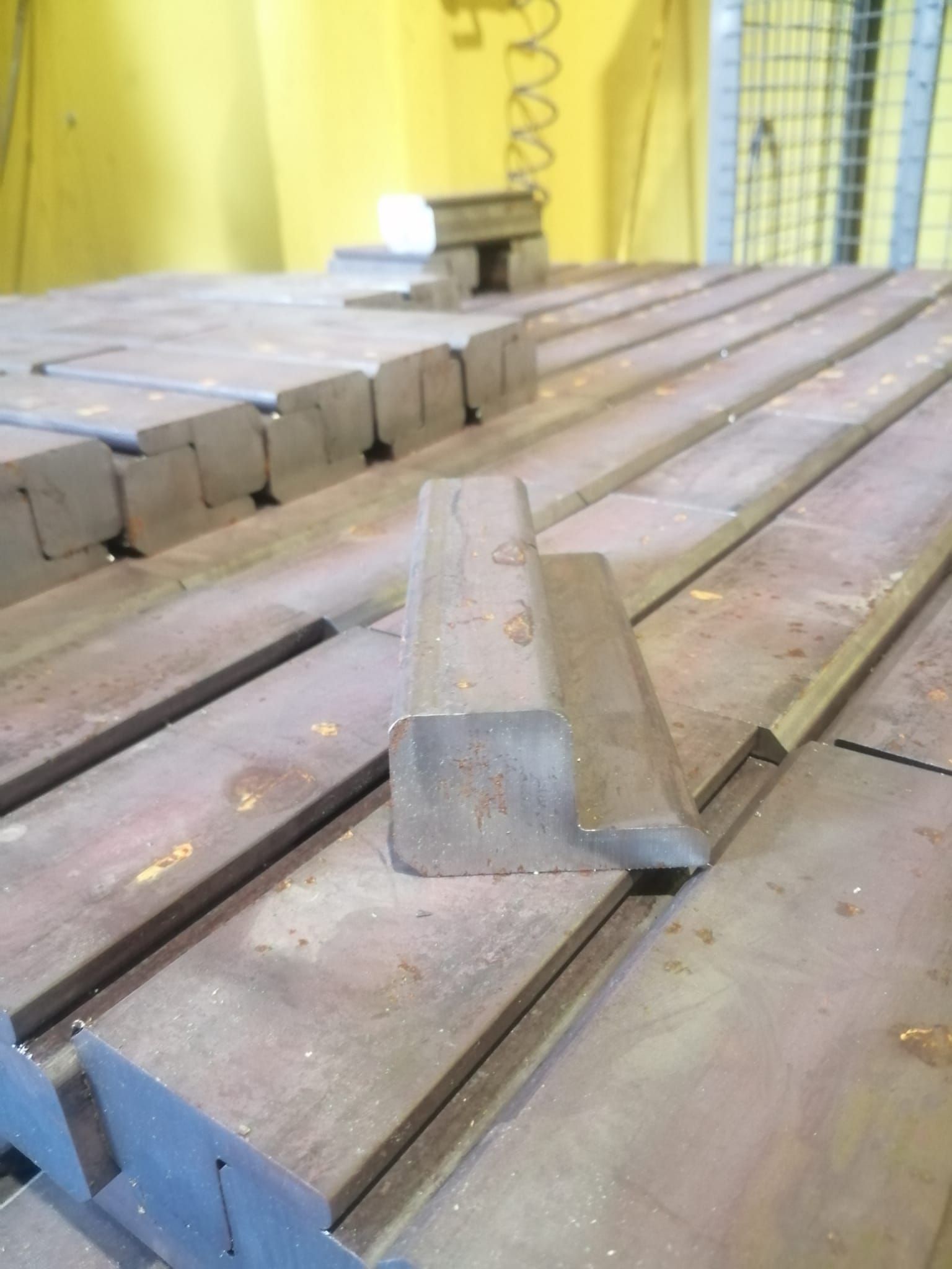 Un blocco metallico a forma di L è posizionato su un basamento industriale a più livelli in un'area di lavoro con una parete gialla sullo sfondo.