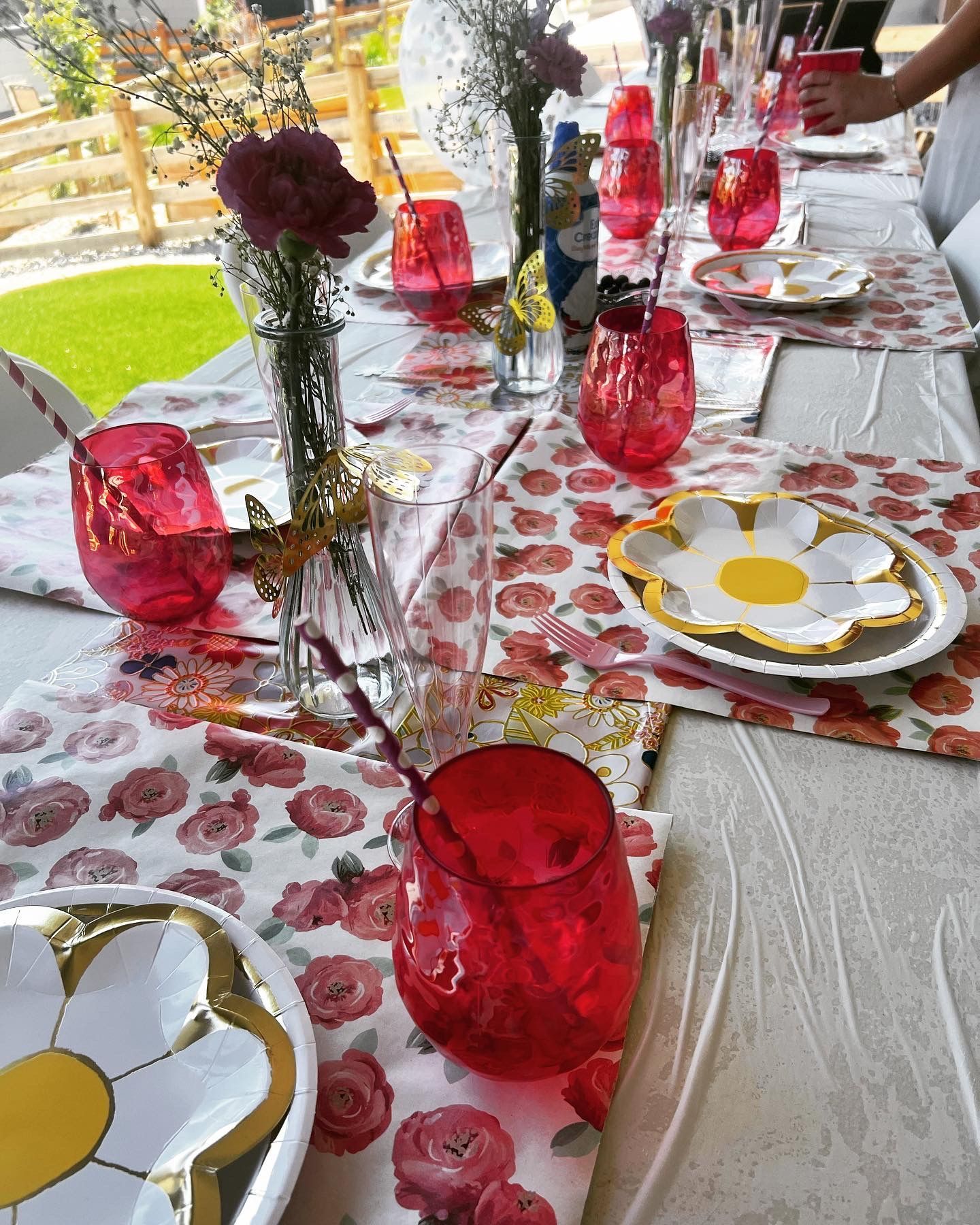 Outdoor table setting with floral tablecloth, pink glasses, daisy plates, and flower centerpieces.