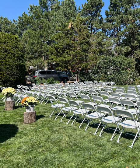 Rows of white folding chairs set up on grass for an outdoor event, with trees in the background.