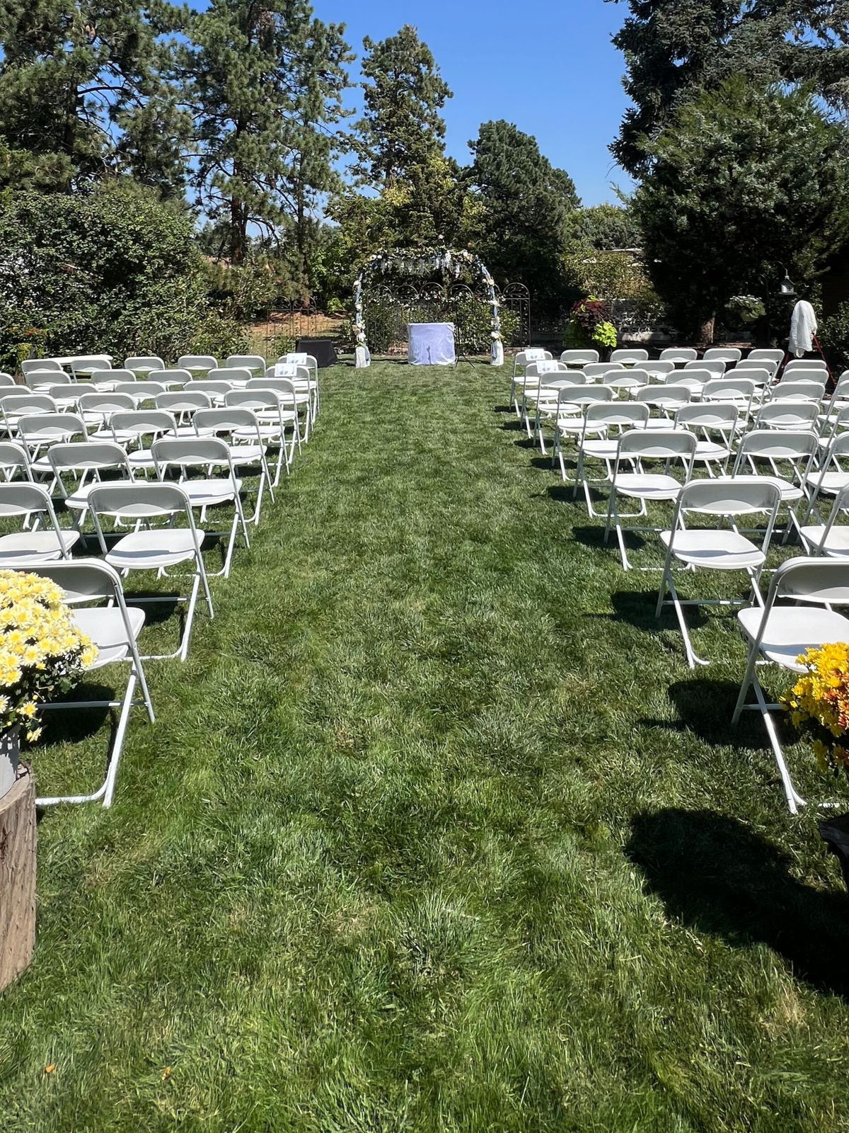 Rows of white folding chairs on green grass lead to an archway, prepared for an outdoor ceremony.