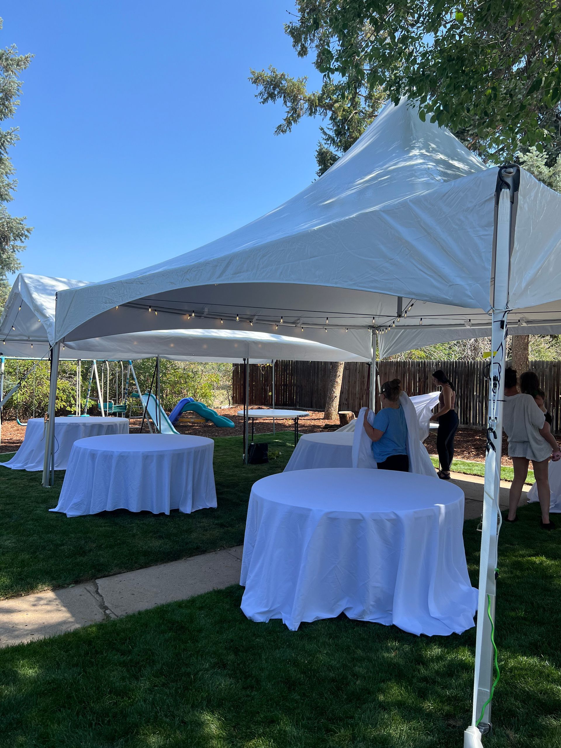 White tents cover round tables in a grassy backyard. People stand nearby on a sunny day.