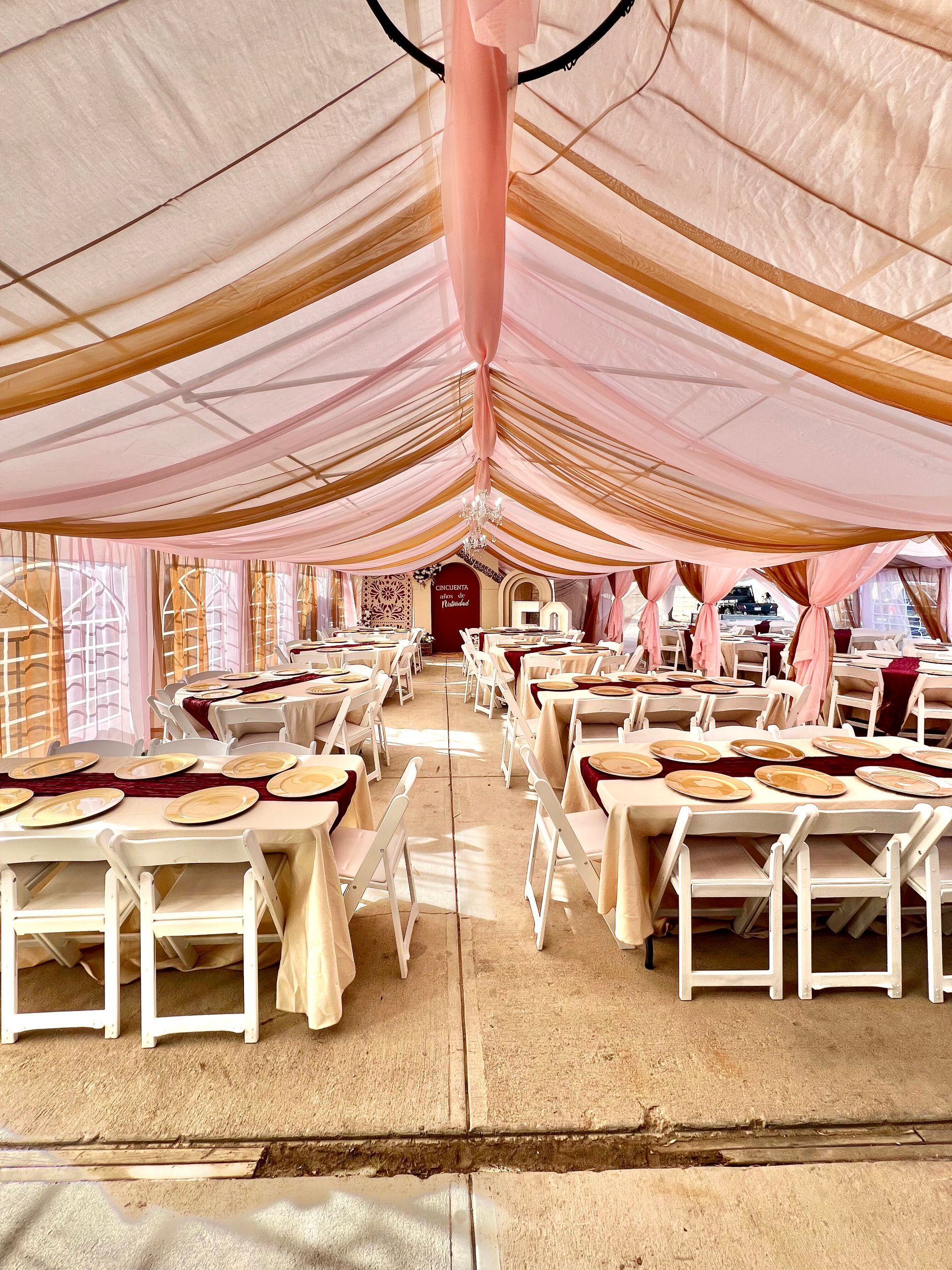 Inside a tent set for a celebration; rows of tables with white chairs and gold/burgundy decor.