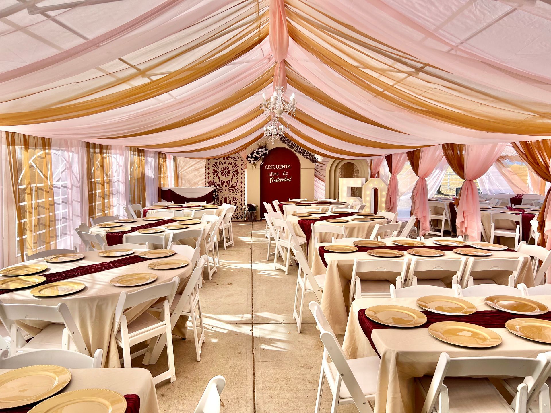 Wedding reception tent with tables set for guests, draped with gold, pink, and burgundy fabric.