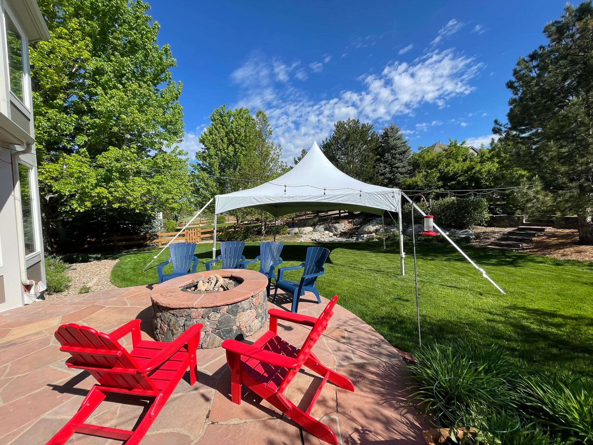 Backyard scene with tent over chairs around a fire pit and red and blue chairs.