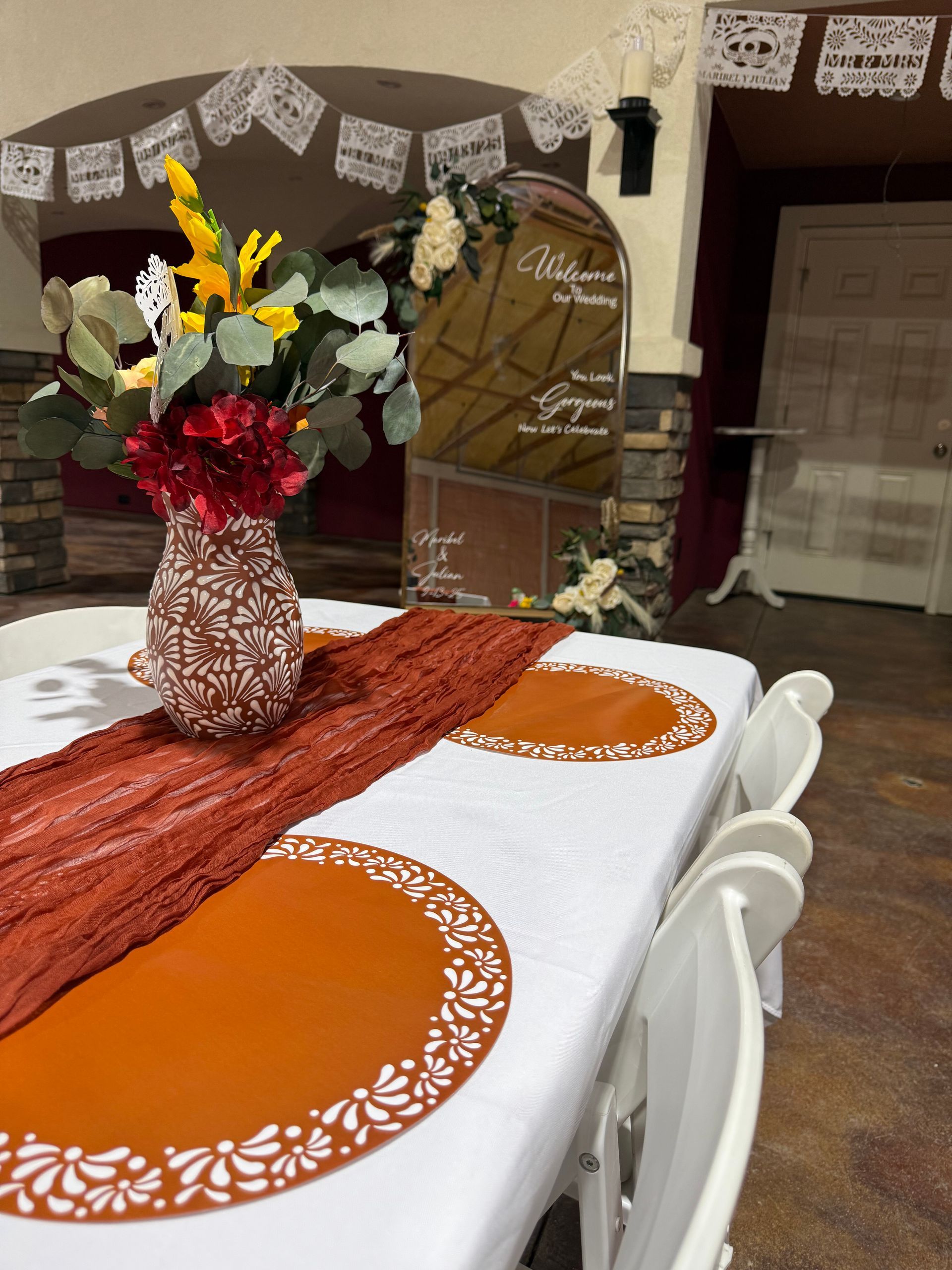Table setting with a floral arrangement, orange runner, placemats, and white chairs in a decorated room.