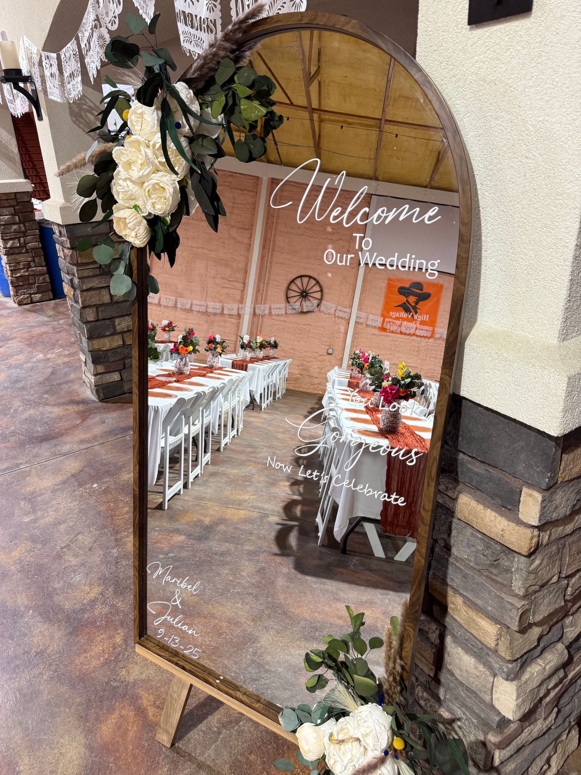 Large mirror with wedding details, arched frame, floral decorations, reflecting wedding reception setup.
