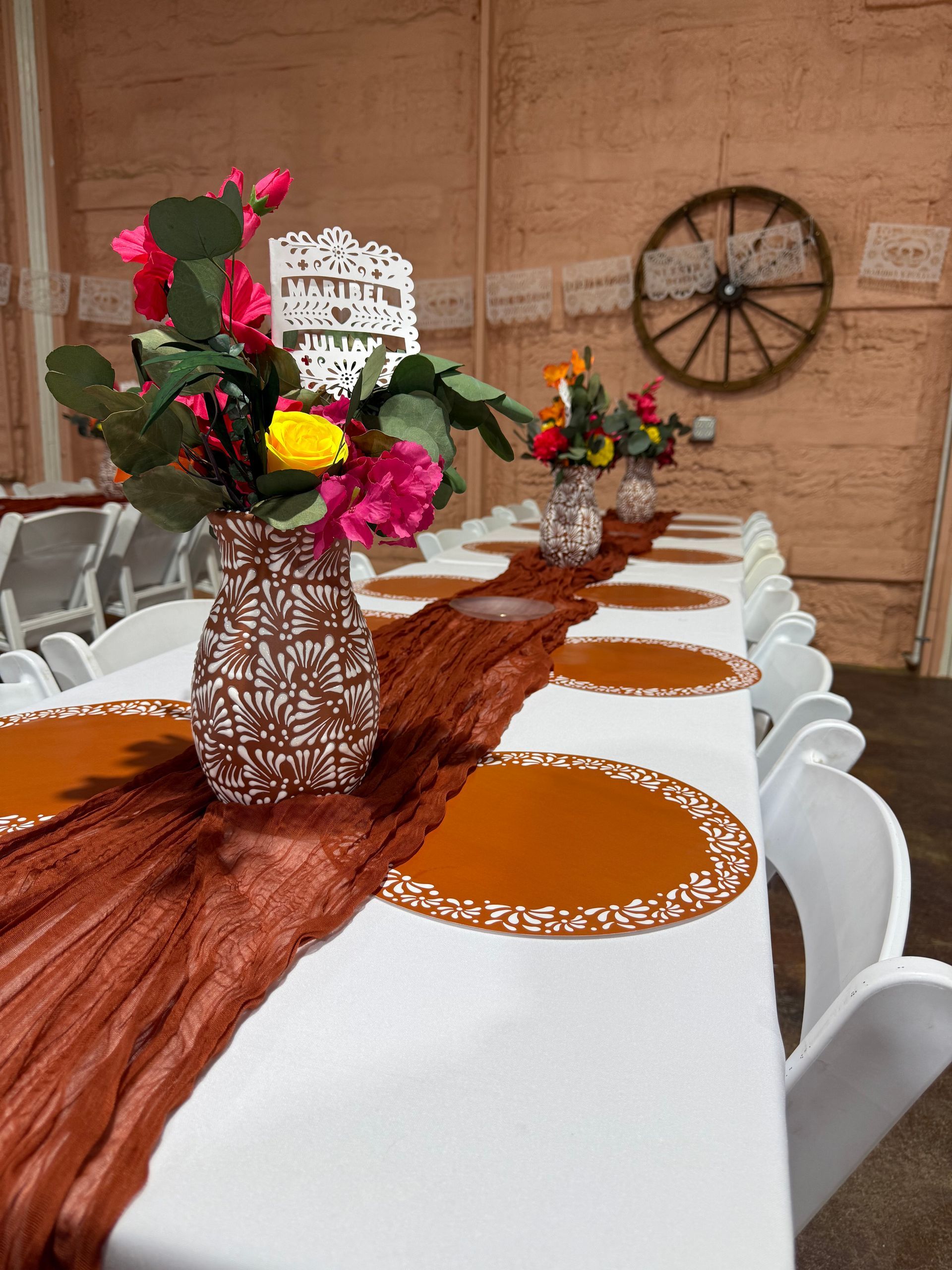 Long table set for a celebration with floral centerpieces, russet runner, and white chairs.