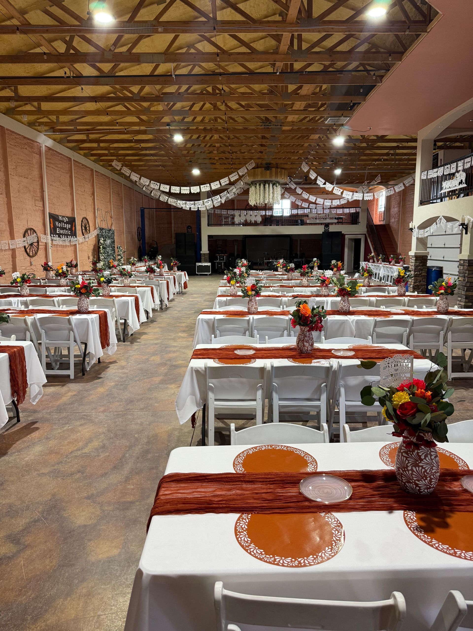 Long banquet hall set with tables and floral centerpieces; terracotta table runners and ceiling decorations.