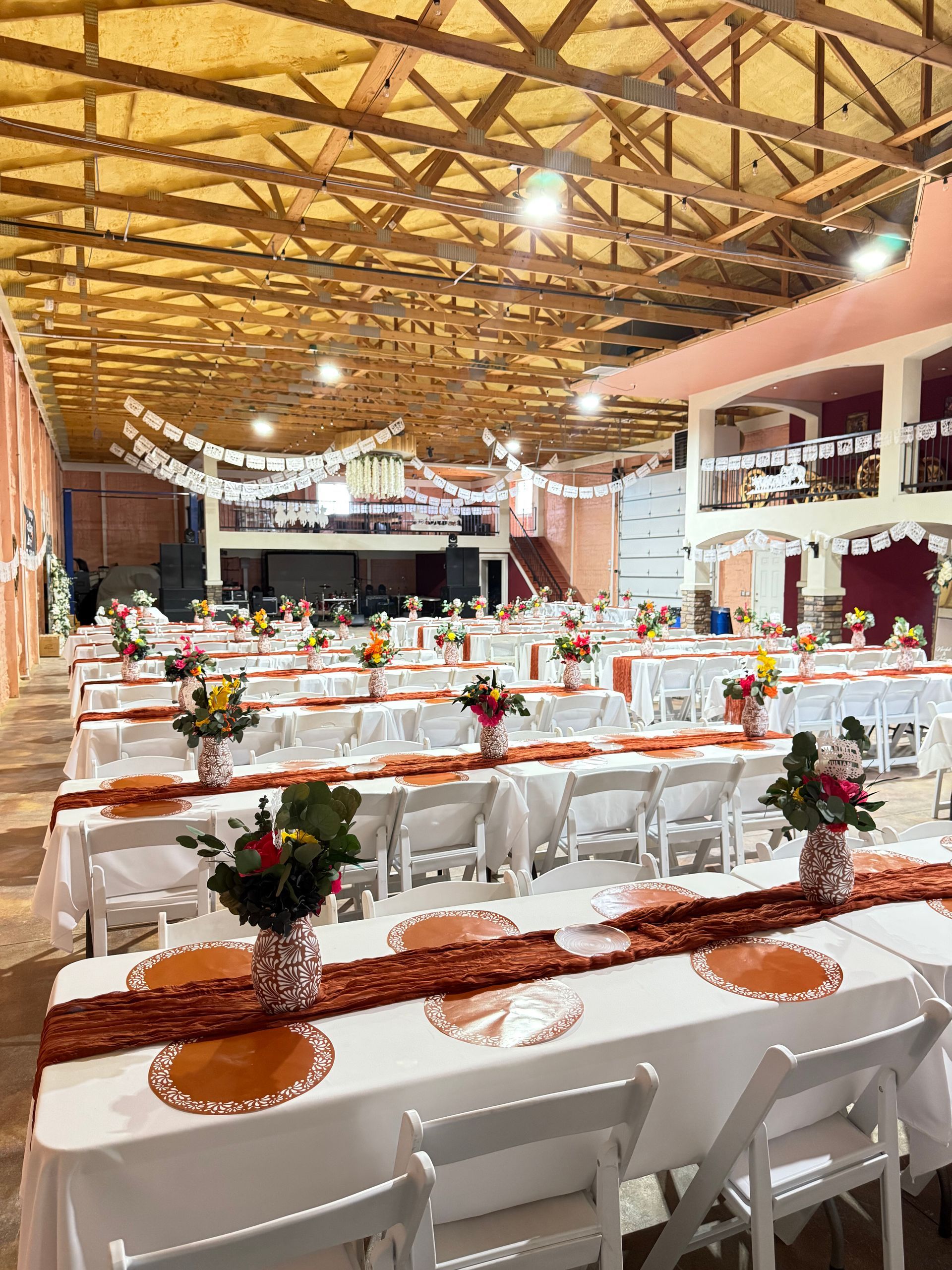 Reception hall set up with white tables, orange runners, and floral centerpieces. String lights and banners hang from the ceiling.