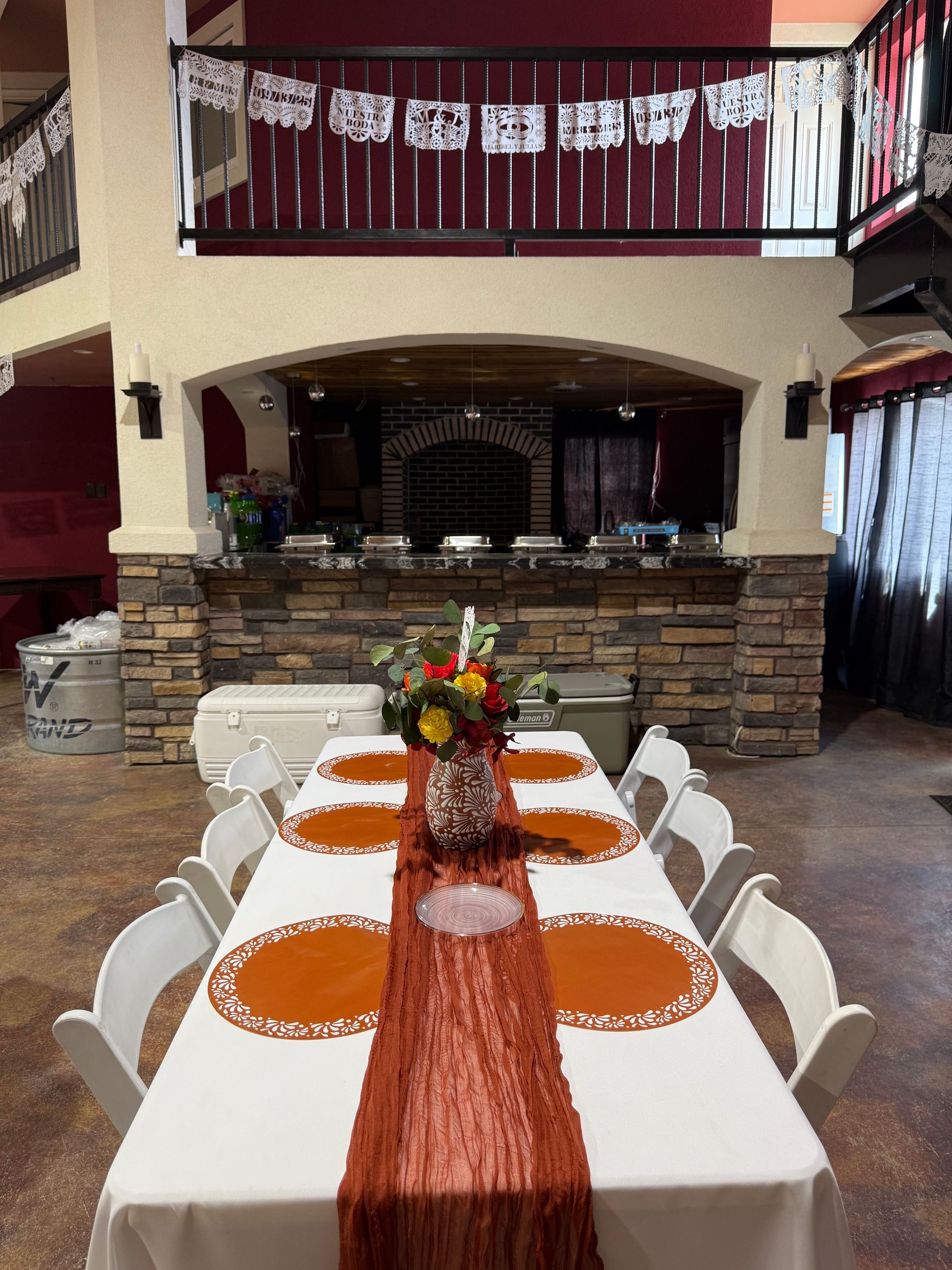 Long table set for a celebration with orange and white decor, brick bar in background, and upper balcony.